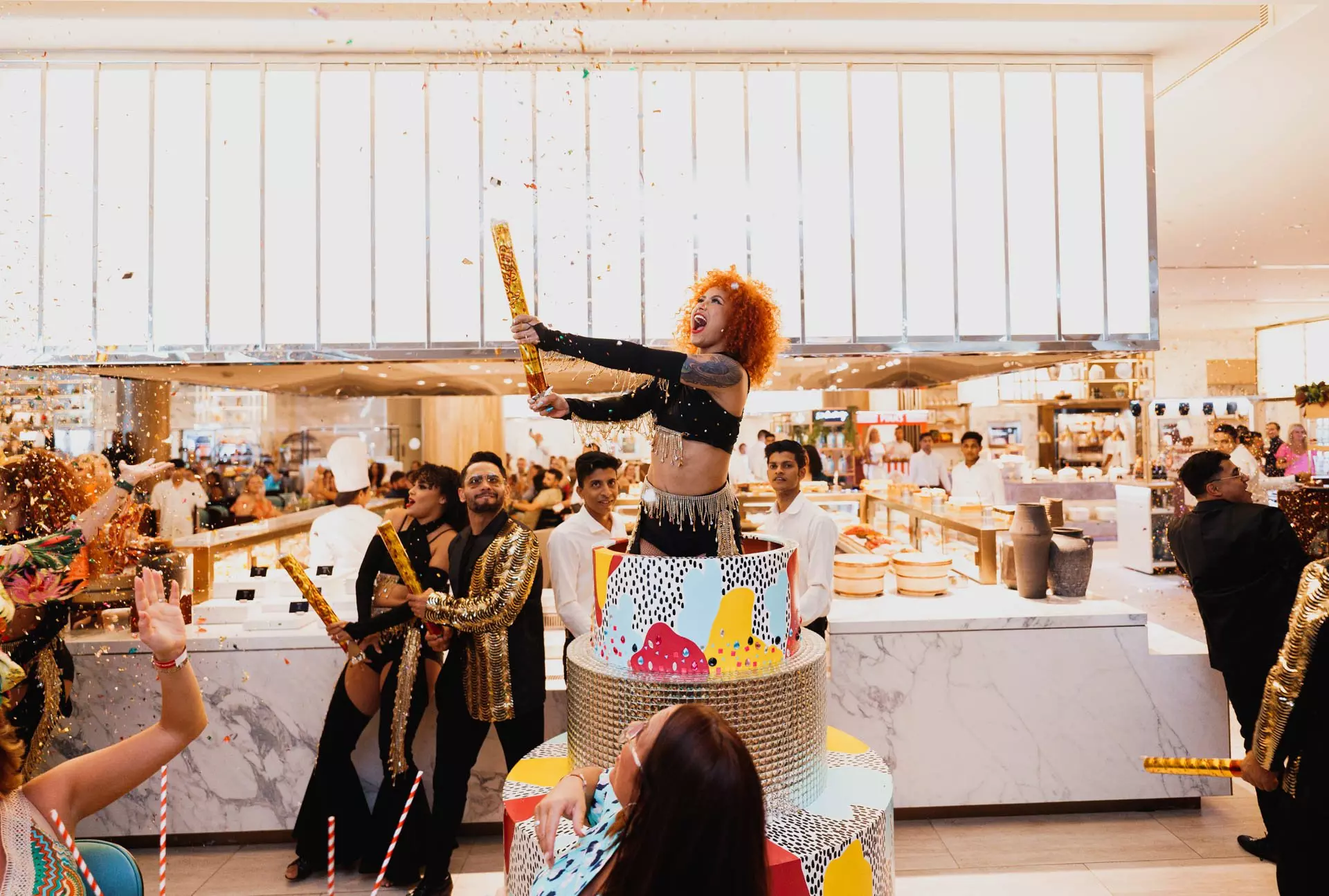 A performer in a sequinned costume fires confetti cannons over a painted cake in the Saffron dining room, with the live cooking stations behind