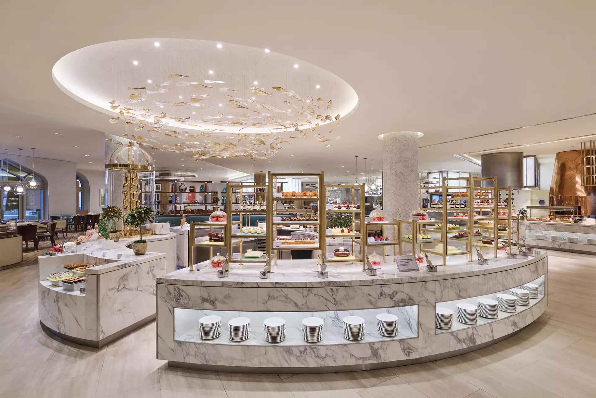 The dessert counter at Saffron: curved marble and gold shelving stacked with cakes and pastries under a chandelier of glass fish