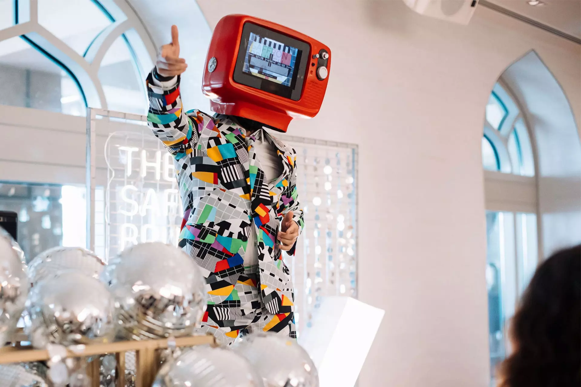 A performer in a patterned suit and a red television helmet above a rack of silver balloons at the Saffron Brunch
